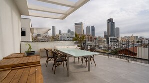 A rooftop terrace with a glass table and chairs, a wooden bench, and a city skyline view. - Luxury Penthouse • Balcony • Parking | HaKerem P2 (680012)