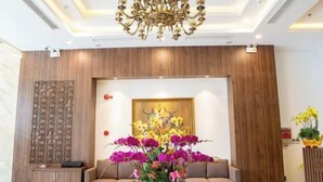 A modern lobby with a sofa, a decorative wall panel, a chandelier, and a floral arrangement. - Minh Quan Hotel - Da Nang Center by Hos (Da Nang)