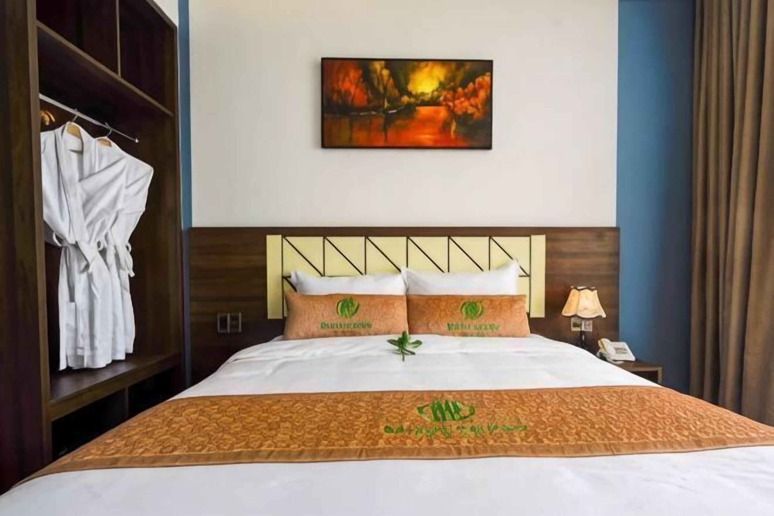 Habitación de hotel con cama, un cuadro en la pared, una bata colgada en un perchero y una mesita de noche con lámpara.