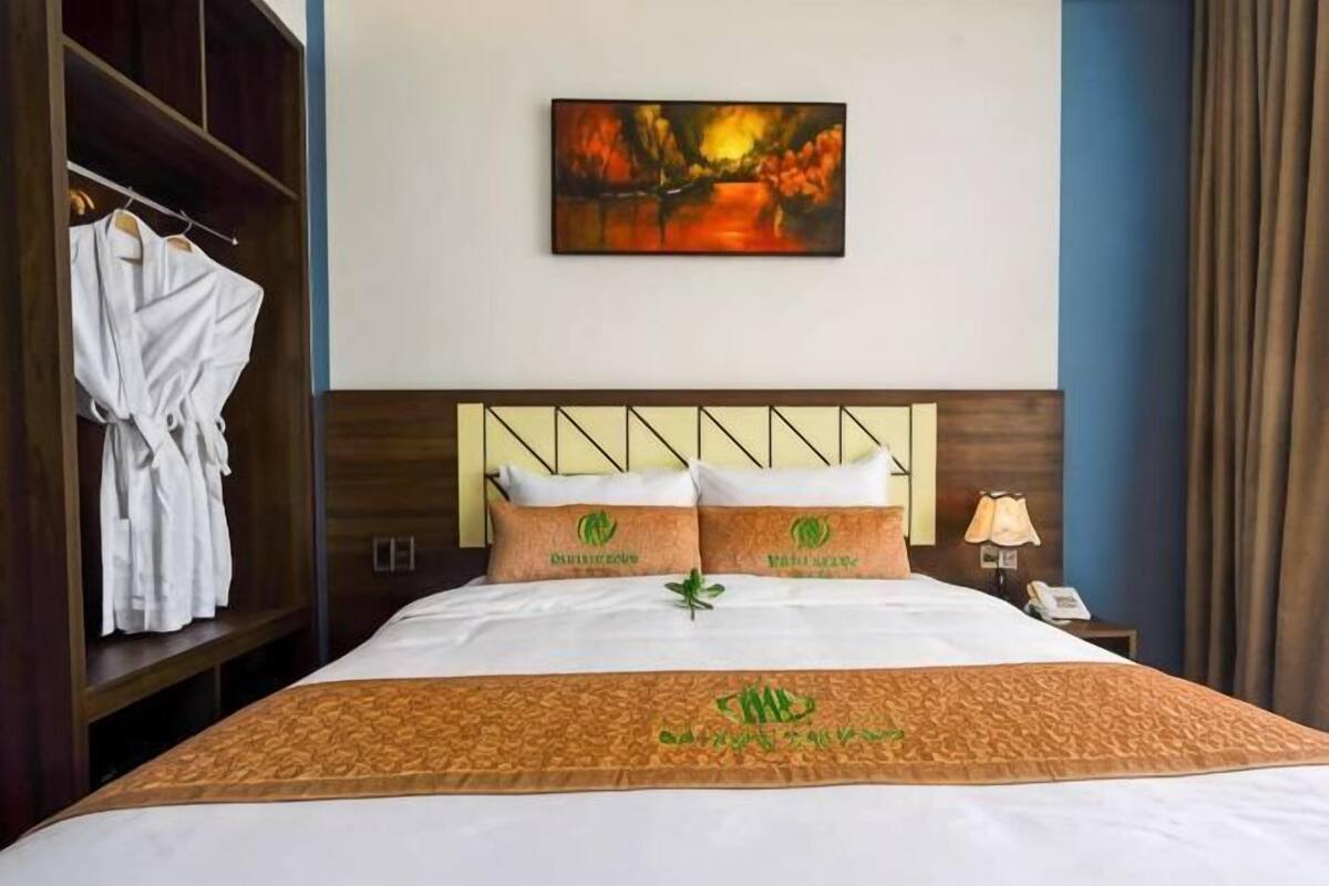 Habitación de hotel con cama, un cuadro en la pared, una bata colgada en un perchero y una mesita de noche con lámpara.
