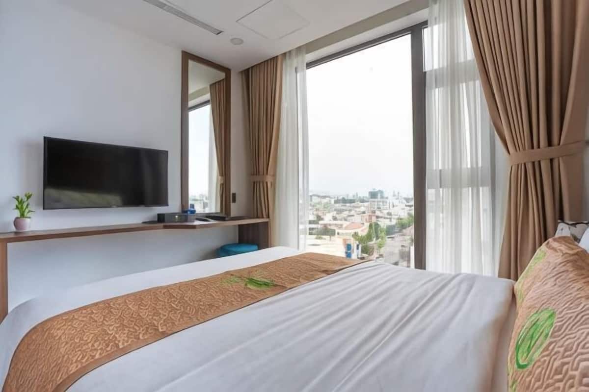 A hotel room with a bed, a TV, a window with curtains, and a view of the city.