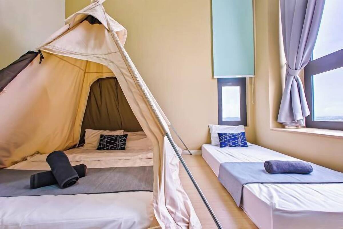 Een kamer met twee bedden, een tentachtige constructie, een raam met gordijnen en een nachtkastje met een lamp.
