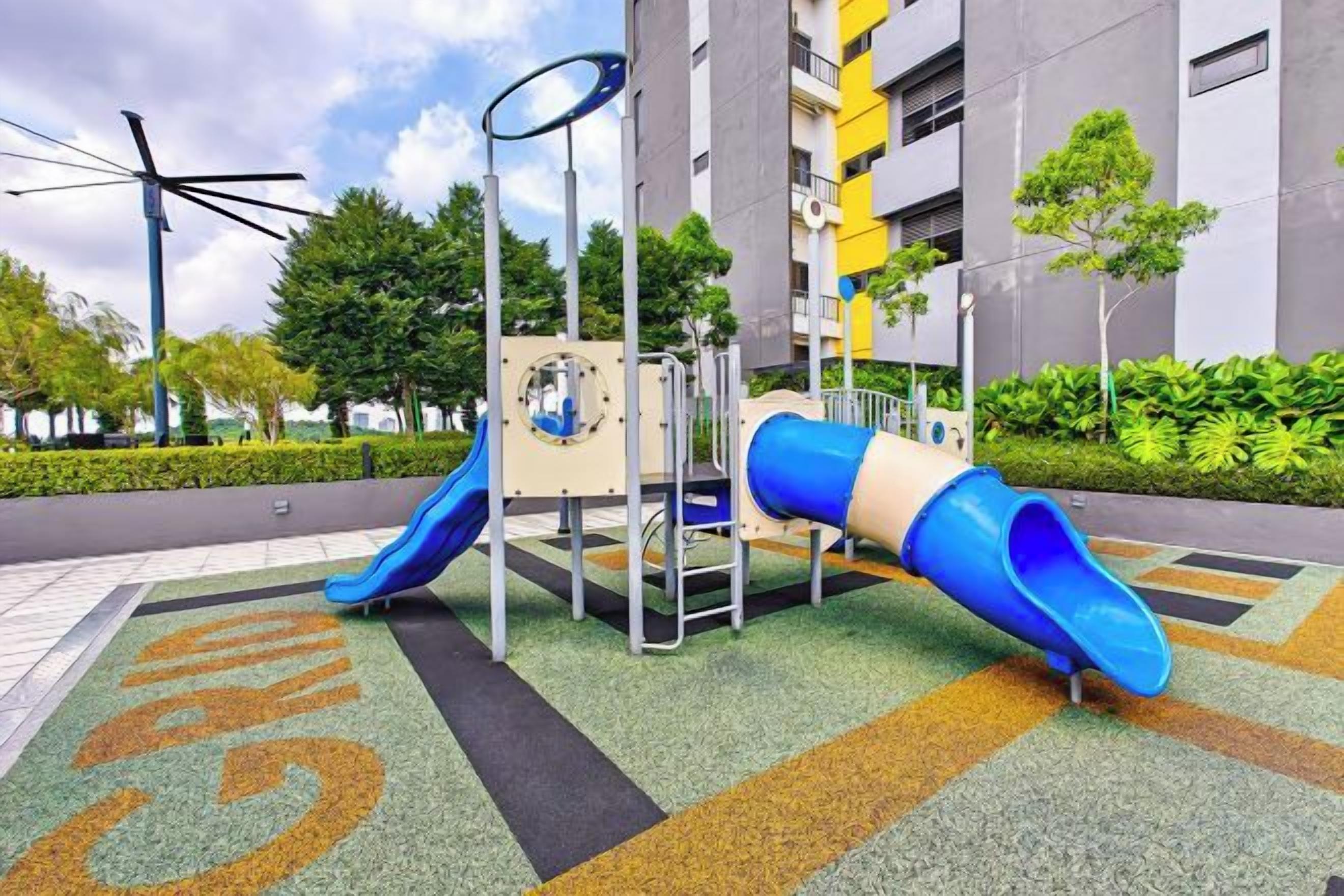 A playground with slides and climbing structures, surrounded by a landscaped area with trees and a building in the background.