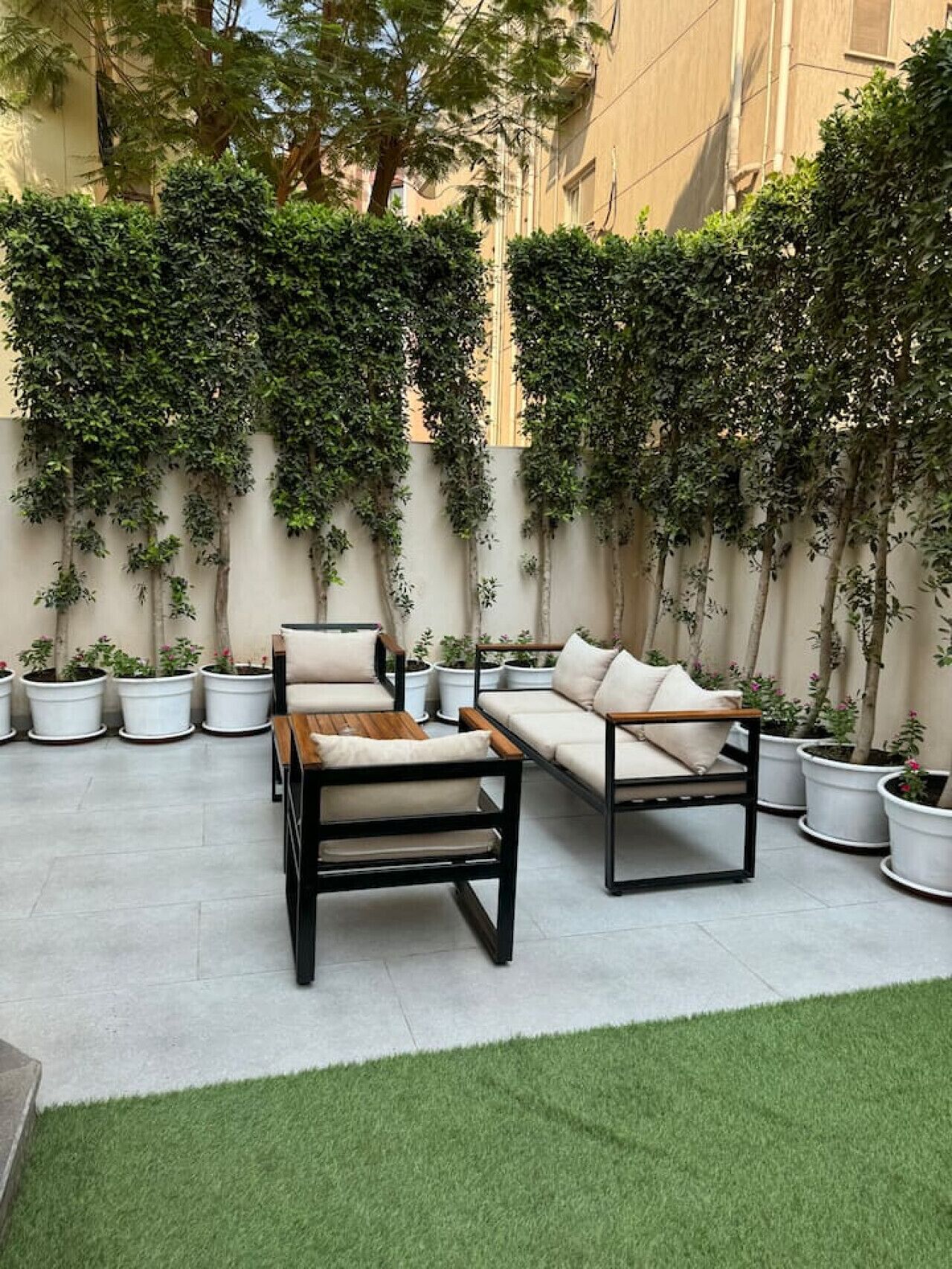 An outdoor seating area with two sofas and a coffee table, surrounded by potted plants and a low hedge.