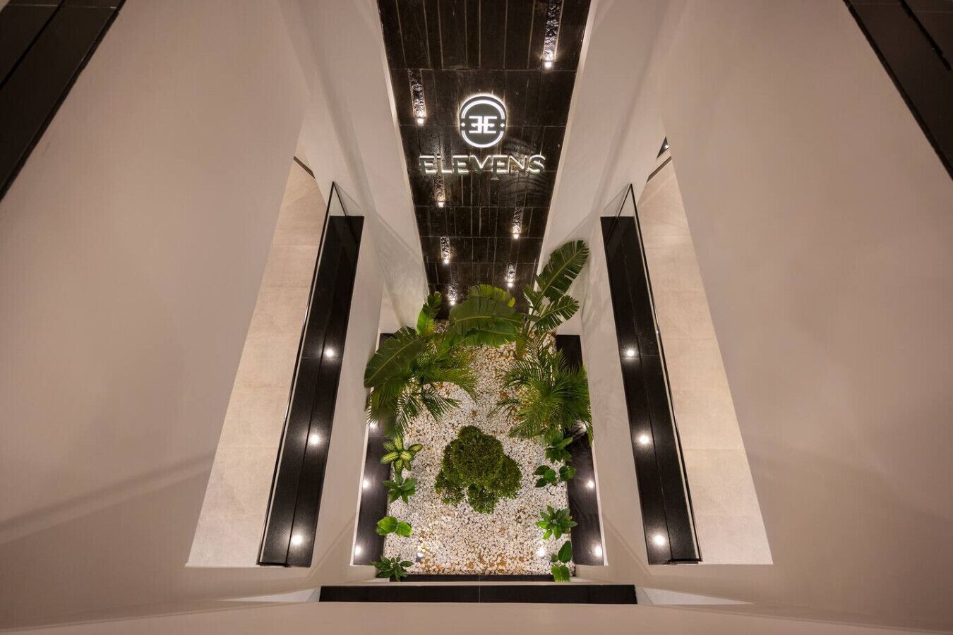 A modern hotel lobby with a central garden area, a prominent sign reading "ELVEN'S", and a sleek, minimalist design.