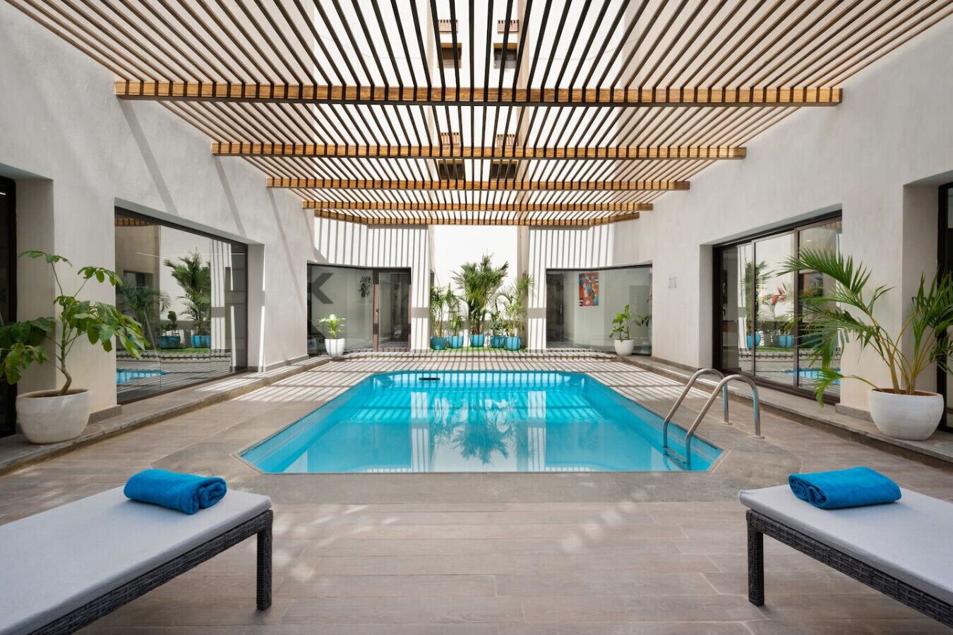 Une piscine intérieure moderne avec un plafond en bois, deux chaises longues avec des serviettes et des plantes en pot disposées autour de la piscine.