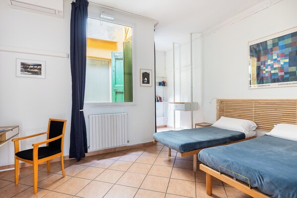 A room with two beds, a chair, a radiator, a window with curtains, and a painting on the wall.