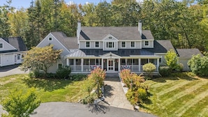 A large, two-story house with a covered porch, surrounded by a well-maintained lawn and trees. - 6 bedrooms! Hot tub/firepit/deck/Pet friendly close to beach! (Kennebunk)