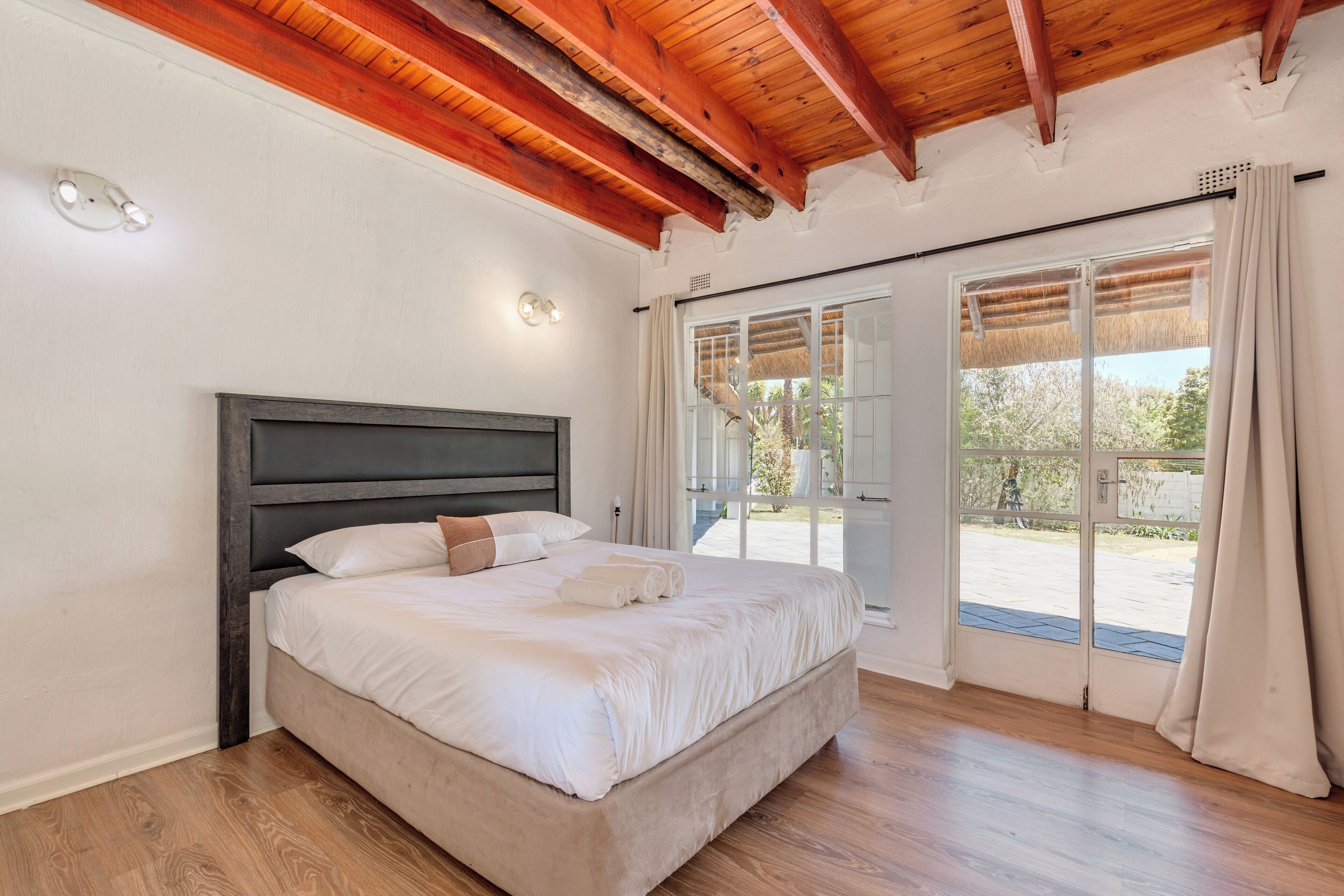 Un dormitorio con una cama grande, vigas de madera en el techo, una puerta corrediza de vidrio al exterior y vistas a la vegetación.