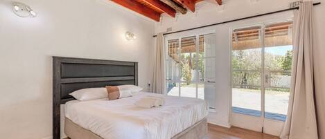 Un dormitorio con una cama grande, vigas de madera en el techo, una puerta corrediza de vidrio al exterior y vistas a la vegetación.