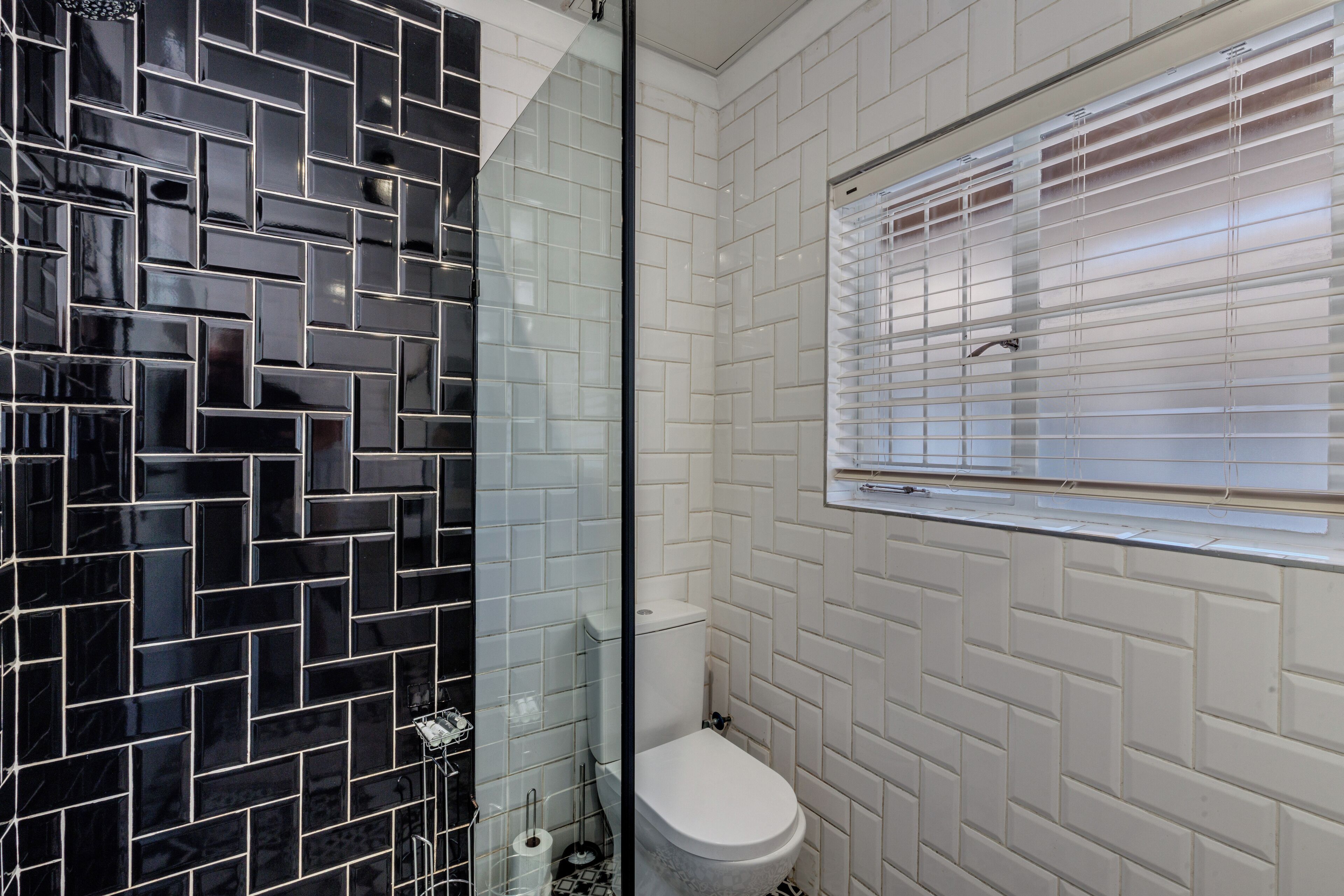A bathroom with black and white tiled walls, a white toilet, and a window with blinds.