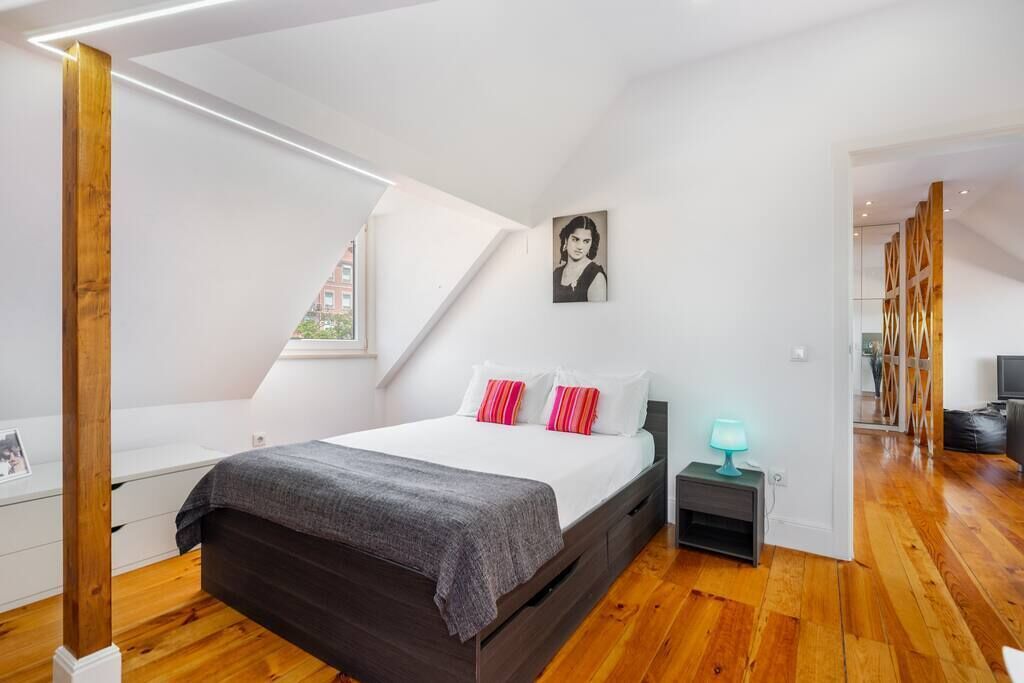 A modern bedroom with a wooden floor, a bed with white and pink pillows, a nightstand with a lamp, and a painting on the wall.