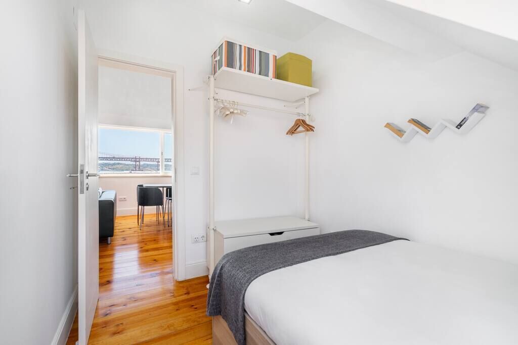Une chambre avec un lit, une armoire blanche, une étagère remplie de livres et une boîte verte, et une vue sur un balcon avec une balustrade.