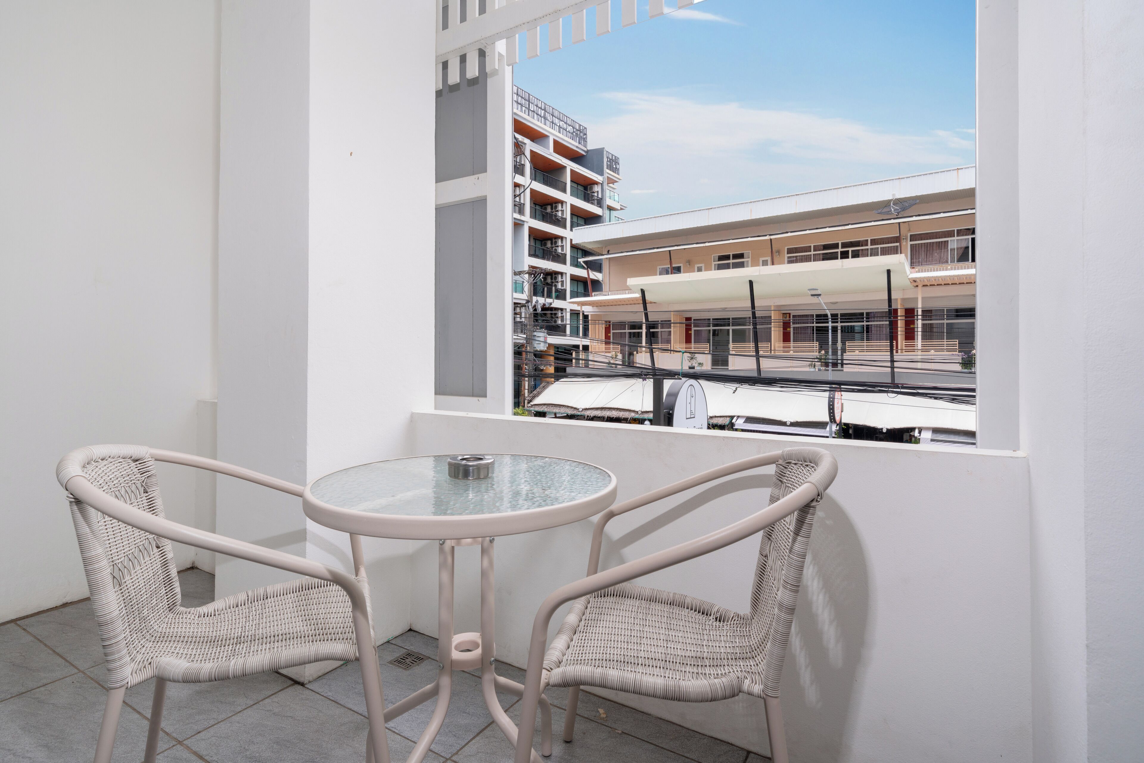 A balcony with two wicker chairs and a glass table, overlooking a building with multiple floors and a covered area.