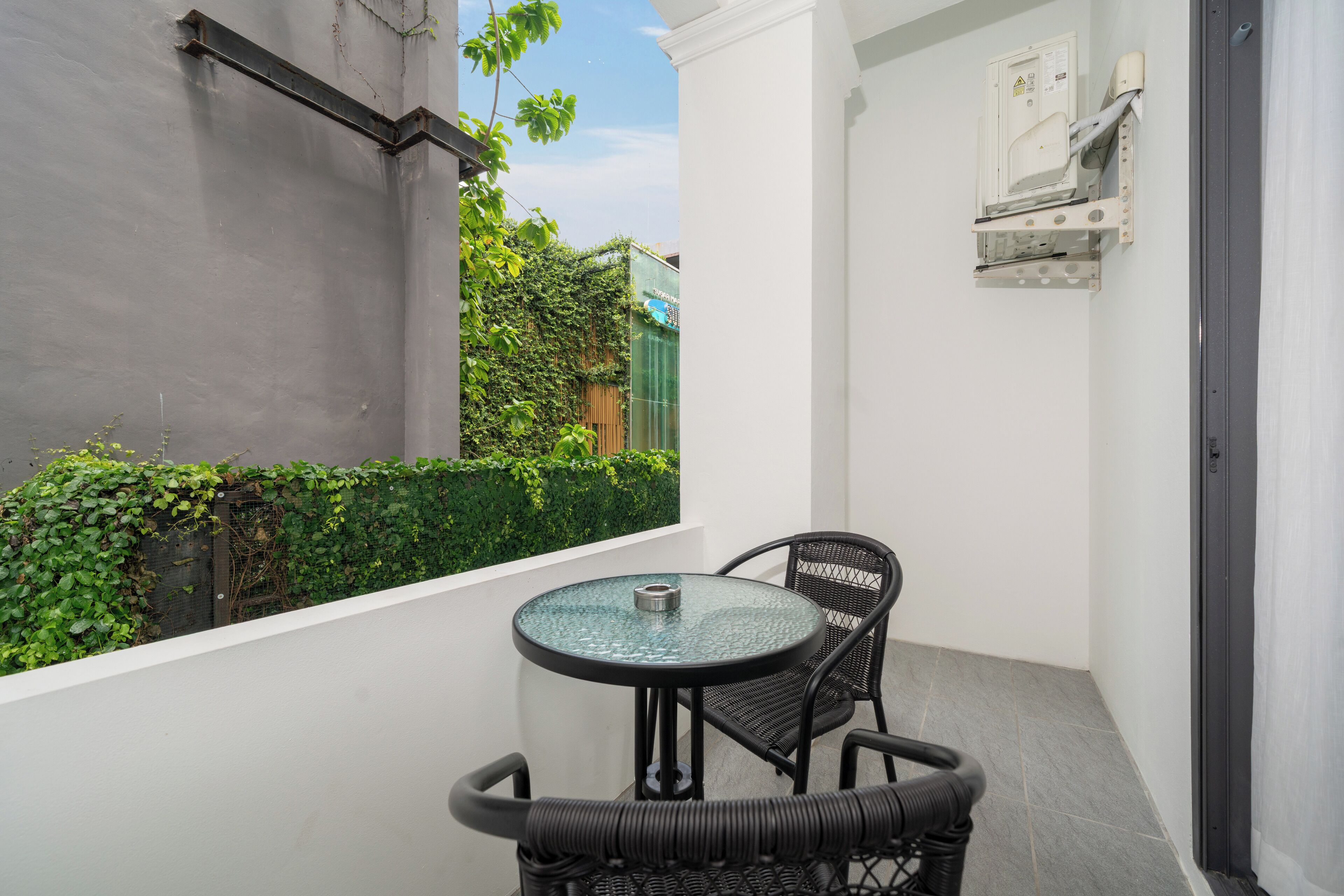 Un petit balcon avec une table ronde en verre et deux chaises en osier, un mur blanc et une haie verte en arrière-plan.