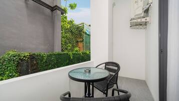 Un petit balcon avec une table ronde en verre et deux chaises en osier, un mur blanc et une haie verte en arrière-plan.