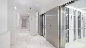 Un corridoio moderno con pavimento in marmo, un ascensore in vetro e una porta con il segnale di allarme antincendio.