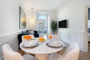 Un coin repas avec une table ronde dressée pour quatre personnes, des murs blancs, un lustre et une vue sur le paysage urbain, visible à travers une porte en arc de cercle.