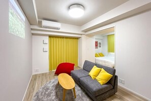 A modern hotel room with a grey sofa, yellow pillows, a wooden side table, and a mirror reflecting the room. - Park Axis Honjo Azumabashi South Residence (Tokyo)
