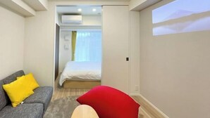 A hotel room with a grey sofa, red and yellow pillows, a bed with white bedding, a large window with curtains, and a wall-mounted projector screen displaying a mountain scene. - Park Axis Honjo Azumabashi South Residence (Tokyo)