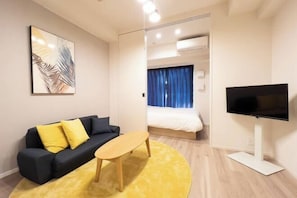 A modern hotel room with a sofa, a small table, a TV, and a bed with a blue curtain. - Park Axis Honjo Azumabashi South Residence (Tokyo)