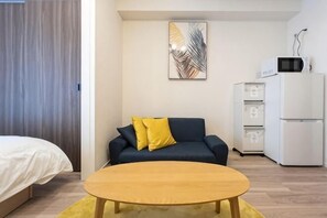 Une chambre moderne comprenant un lit, un canapé avec des coussins jaunes, une table basse en bois, un four à micro-ondes et un réfrigérateur.