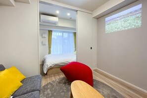 A hotel room with a bed, a gray sofa, a red chair, a wooden table, and a projection screen on the wall. - Park Axis Honjo Azumabashi South Residence (Tokyo)