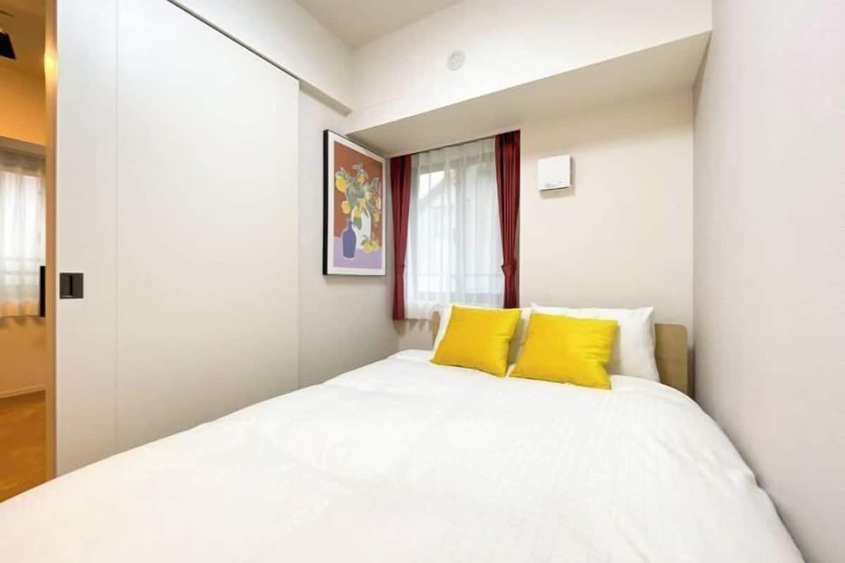Habitació d'hotel amb un llit, parets blanques, un quadre i una finestra amb cortines vermelles.