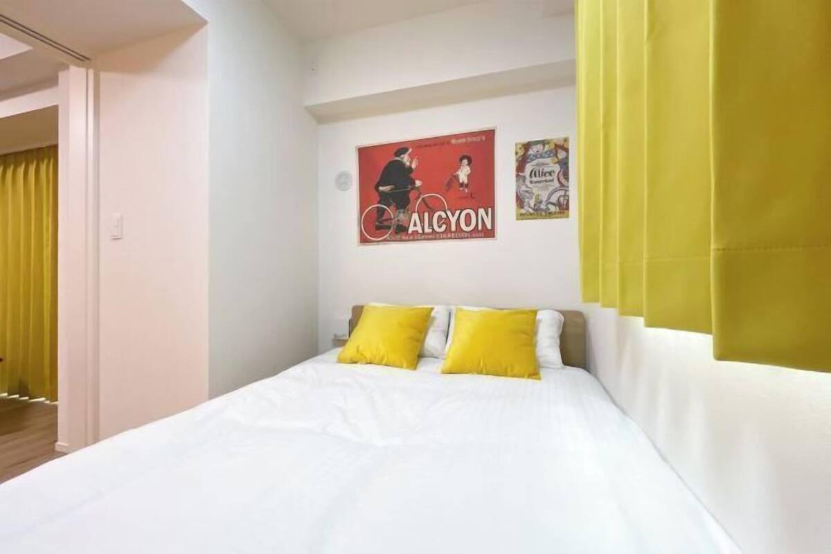 Habitació d'hotel amb un llit, almoinades grogues, un cartell a la paret i una cortina groga.