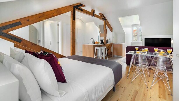 A modern loft bedroom with a large bed, wooden beams, a kitchenette, and a dining area with clear chairs.