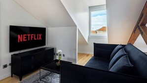 Ein modernes Wohnzimmer mit einem schwarzen Sofa, einem Couchtisch, einer schwarzen TV-Konsole mit Netflix-Logo und einem Fenster mit Blick auf Gebäude.