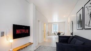 A modern living room with a black sofa, a mounted TV displaying the Netflix logo, a wooden shelf, and a patterned rug. - Lisbon Prime Aparts - BeatoHub (Lisboa)