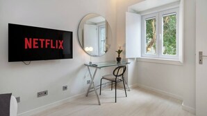 A modern room with a large flat-screen TV mounted on the wall, a round mirror, a glass desk with a chair, and a window with white frames. - Lisbon Prime Aparts - BeatoHub (Lisboa)