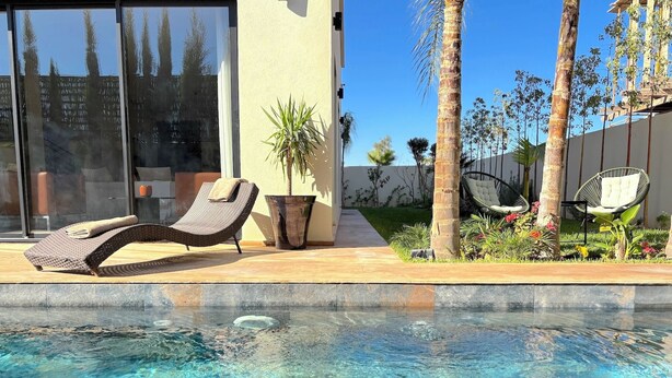 Un espace piscine avec une eau claire, une chaise longue, une plante en pot et un hamac.