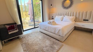 Une chambre moderne avec un grand lit, une porte-fenêtre coulissante donnant sur un balcon, une table de chevet blanche et un miroir décoratif mural.