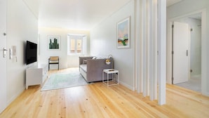 Un salon moderne avec un plancher en bois, un canapé gris, une petite table, un téléviseur et une porte donnant sur une autre pièce.