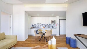 A modern living room with a sofa, a dining area with chairs, a kitchen with white cabinets, and a TV mounted on the wall. - Alcântara Premium 2-bed | Wi-Fi | Lx Factory (Lisboa)