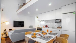 A modern kitchen with white cabinets, a built-in microwave, and a refrigerator. There is a dining table with chairs and a sofa in the living area.