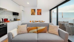 A modern living space with a grey sofa, striped cushions, a kitchen with white cabinets, and a balcony with a view.