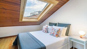 Een slaapkamer met een schuine plafond, een dakraam, een bed met een blauwe spreideken en een nachtkastje met een lamp.