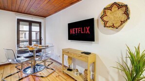 Een moderne woonkamer met een glazen eettafel, een houten console en een televisie aan de muur, plus een grote gevlochten mand.