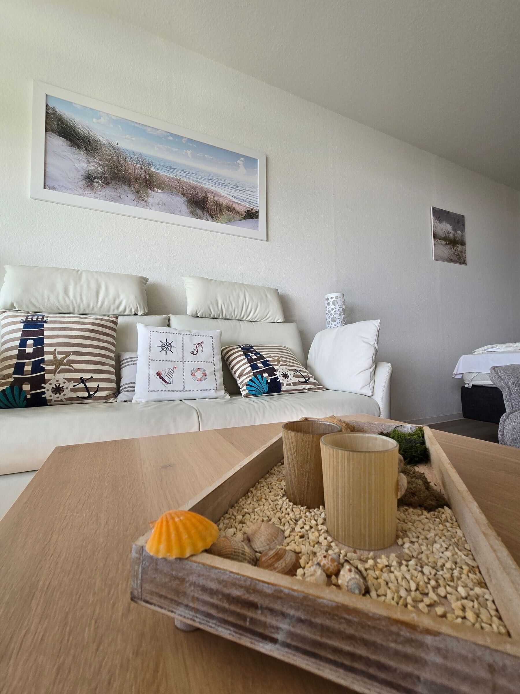 Ein modernes Wohnzimmer mit einem beigefarbenen Sofa, dekorativen Kissen, einem Couchtisch aus Holz mit Meeresschalen und einem Strandbild an der Wand.