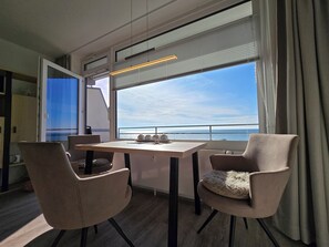 Un espace de repas moderne avec une table et des chaises, de grandes fenêtres offrant une vue sur la mer et un luminaire au plafond.