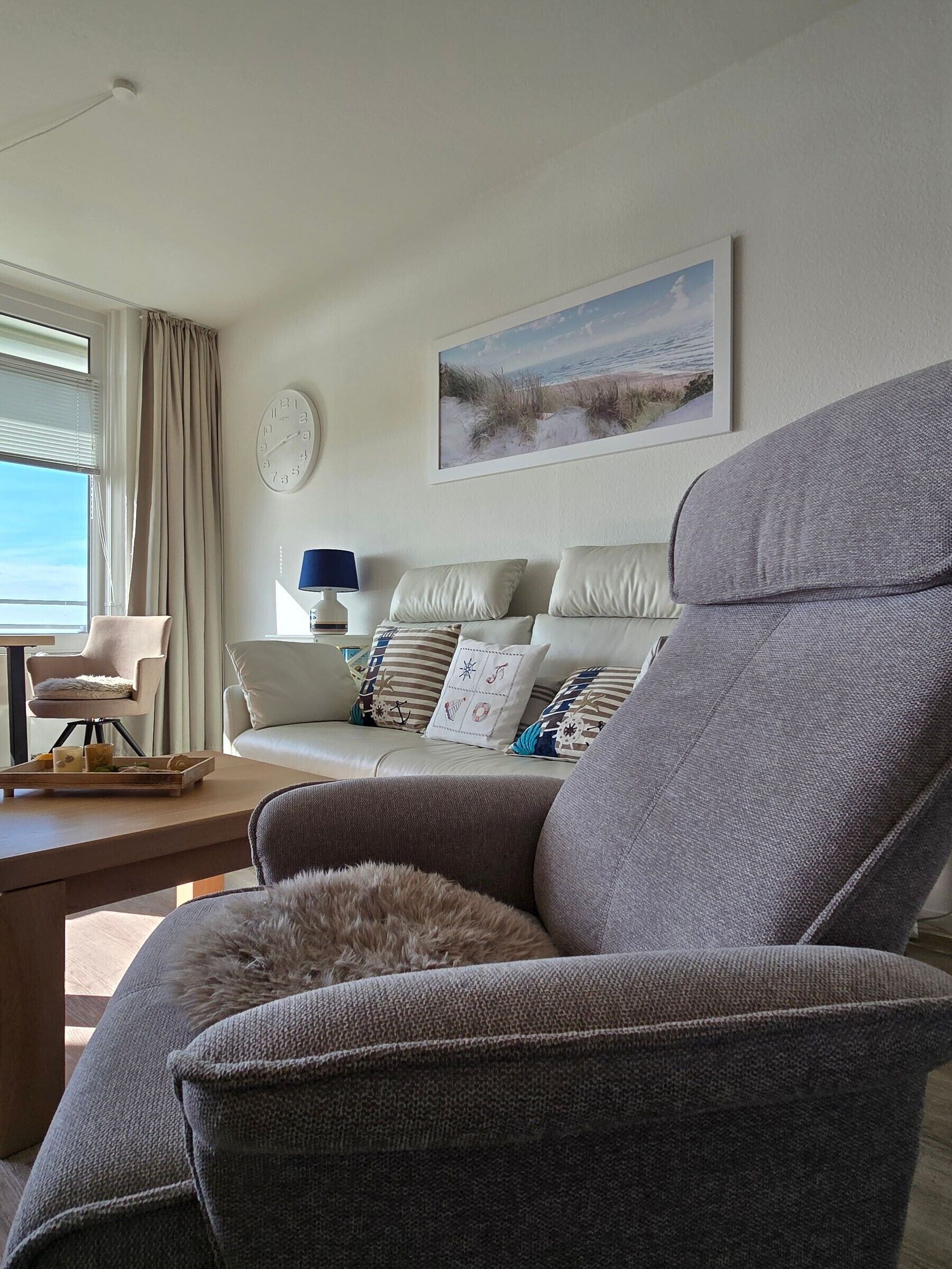 Ein modernes Wohnzimmer mit einem grauen Sessel, einer Couch mit gemusterten Kissen, einem Beistelltisch mit Tablett, einer Lampe und einer Wanduhr.
