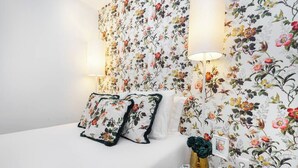 A bedroom with floral wallpaper, a bed with matching pillows, a bedside table with a vase and a lamp.