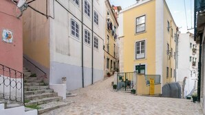A narrow cobblestone street flanked by buildings with balconies and a staircase leading to an upper level.