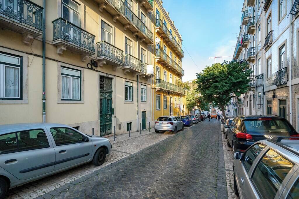 Une ruelle étroite pavée, bordée d’immeubles à plusieurs étages, de voitures garées et d’un arbre qui offre de l’ombre.