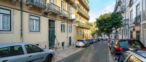 Une ruelle étroite pavée, bordée d’immeubles à plusieurs étages, de voitures garées et d’un arbre qui offre de l’ombre.
