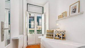 A bedroom with a white bed, a small white table, and a window with a view of buildings.
