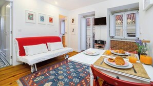 A compact living space with a red sofa, a dining table with breakfast, and a TV mounted on the wall. - Paz 2 | Tram28 | Wi-Fi | AC | Bairro Alto (Lisboa)